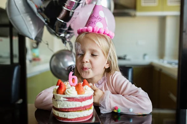 Sarı saçlı, mum üfleyen, 6 yaşındaki doğum gününü kutlayan, şık bir elbise giyen, heyecanlı ifadeler takınan oldukça neşeli küçük bir kızın kapalı mekândaki fotoğrafı. Mutlu çocukluk kavramı — Stok fotoğraf