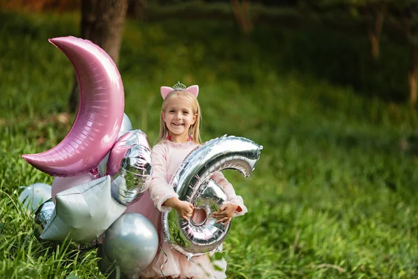 Venkovní záběr pěkné radostné holčičky s blond vlasy slaví 6 let staré narozeniny s balónky, nosit módní šaty, mají vzrušené výrazy. Veselé vzpomínky z dětství — Stock fotografie