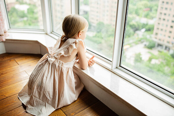 Stylish little girl with blonde hair sitting at home near window during coronavirus covid-19 self isolation. High quality photo
