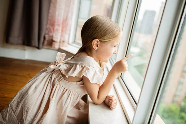 Sarı saçlı şık küçük kız Coronavirus covid-19 izolasyonu sırasında pencerenin yanında oturuyor. — Stok fotoğraf