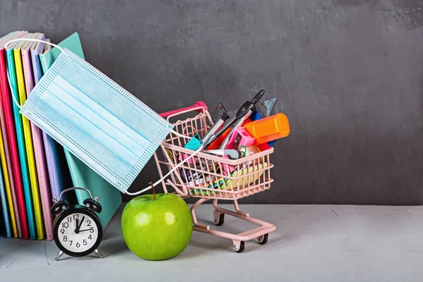 Ritorno a scuola con carrello della spesa e maschera medica. Raccolta di materiale scolastico. Istruzione durante la pandemia di coronavirus covid-19. Copia spazio, mockup, modello. — Foto Stock