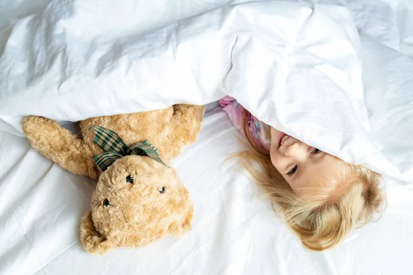 Nettes kleines Mädchen mit blonden Haaren auf dem Bett liegend mit Stoffteddy. Glückliche Kindheit. Bleiben Sie während des Coronavirus-Covid-19-Pandemie-Quarantäne-Konzepts zu Hause. — Stockfoto