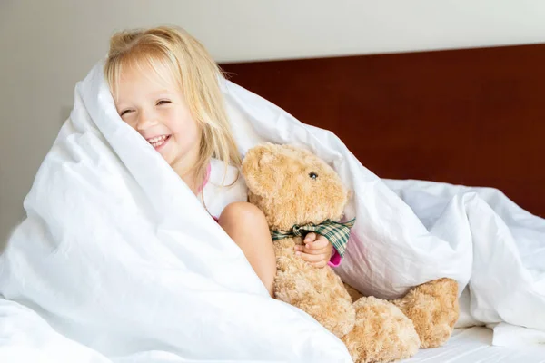 Doldurulmuş oyuncak ayıyla yatakta oturan sarı saçlı tatlı küçük kız. Mutlu bir çocukluk. Coronavirus covid-19 salgın karantina konsepti sırasında evde kal. — Stok fotoğraf
