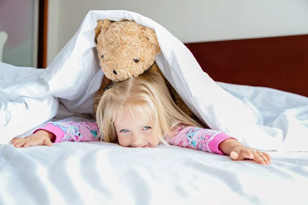 Oyuncak ayıyla yatakta yatan sarı saçlı tatlı küçük kız. Mutlu bir çocukluk. Coronavirus covid-19 salgın karantina konsepti sırasında evde kal. — Stok fotoğraf