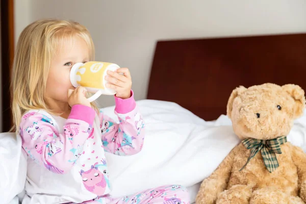 Schattig klein meisje met blond haar op bed en melk drinkend. Gelukkige jeugd. Blijf thuis tijdens het coronavirus covid-19 pandemische quarantaineconcept. — Stockfoto