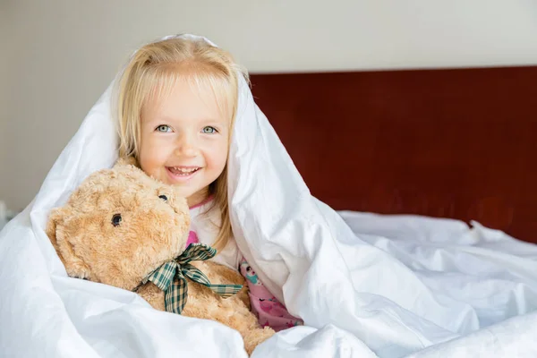 Doldurulmuş oyuncak ayıyla yatakta oturan sarı saçlı tatlı küçük kız. Mutlu bir çocukluk. Coronavirus covid-19 salgın karantina konsepti sırasında evde kal. — Stok fotoğraf