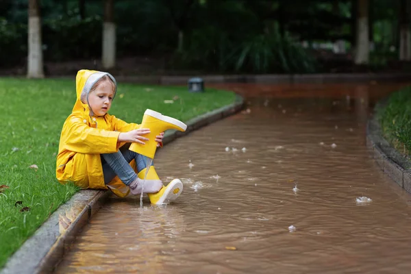 Cute little girl in yellow raincoat and rubber boots walking outdoor during rain. Bad weather, summer tropical storm, autumn fashion concept. — Stock Photo, Image