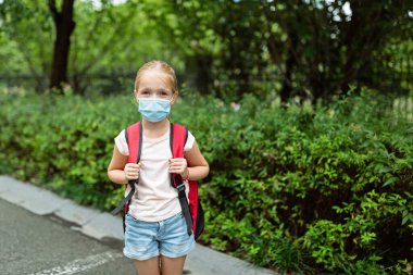 Koronavirüs salgını sırasında yüz maskesi takan okul çocuğu. Sarışın kız, karantina ve tecritten sonra okula geri dönüyor. Coronavirus önleme için tıbbi maskeli bir çocuk. Yeni normal