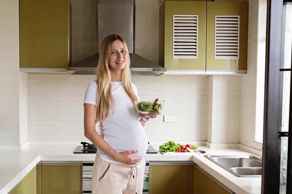 Happy young pregnant woman eating vegetable salad at home. Healthy nutrition and pregnancy concept. Expecting mother holding bowl with fresh cucumbers and tomatoes. High quality photo