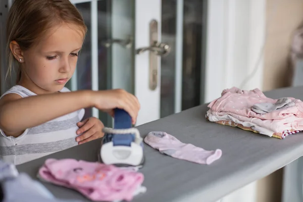 Cute little girl with blonde hair is leaning on ironing clothes on board at home. High quality photo