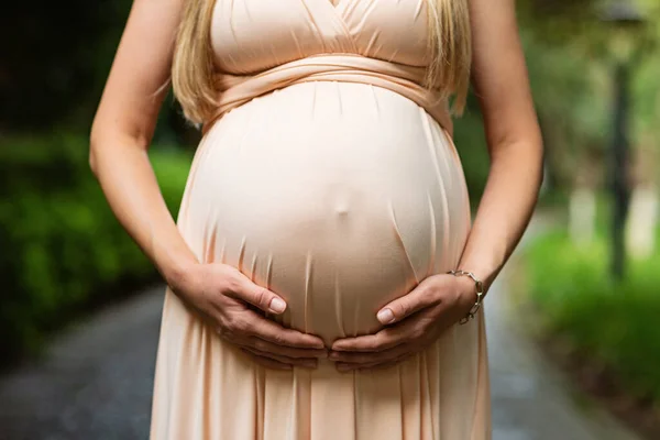 Gece elbisesi giymiş hamile bir kadın yaz parkında karnının üstünde el ele tutuşuyor. Hamilelik, annelik, hazırlık ve beklenti konsepti. Hamileliğin güzel ve hassas fotoğrafı.. — Stok fotoğraf
