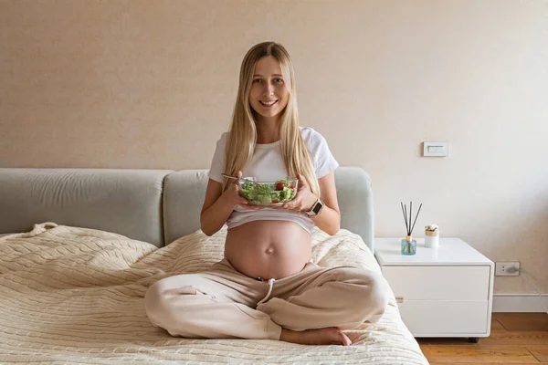 Feliz joven embarazada comiendo ensalada de verduras en casa. Concepto de nutrición saludable y embarazo. Esperando madre sosteniendo tazón con pepinos frescos y tomates —  Fotos de Stock