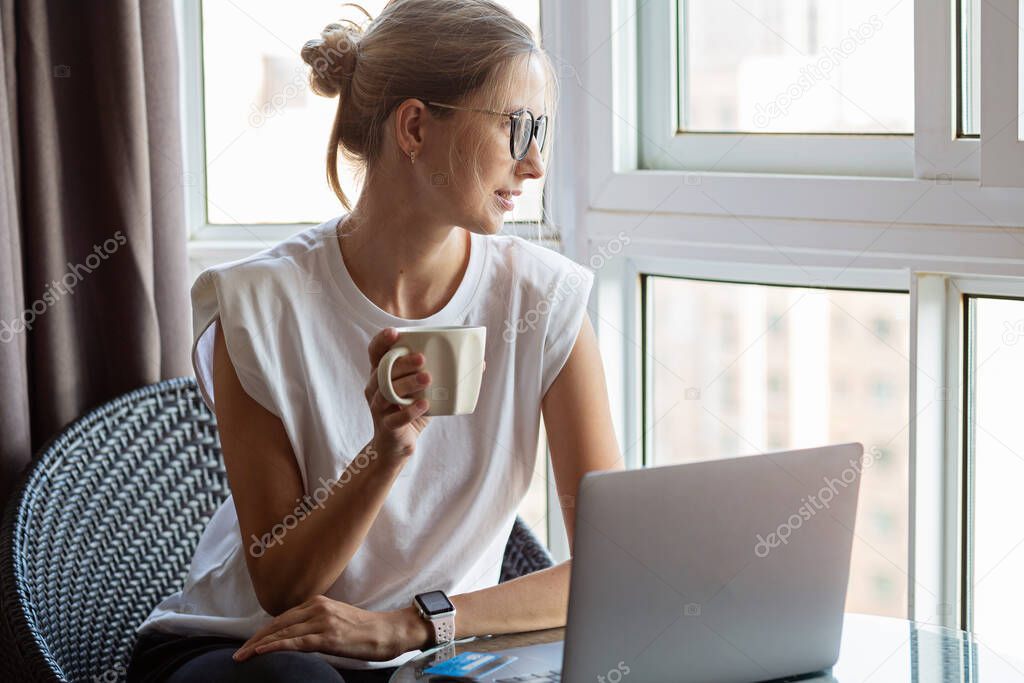 Young woman wearing smartwatch, drinking hot coffee and using laptop. Female working on computer pc at home during covid-19 coronavirus quarantine. Stay home, social distance concept. High quality photo