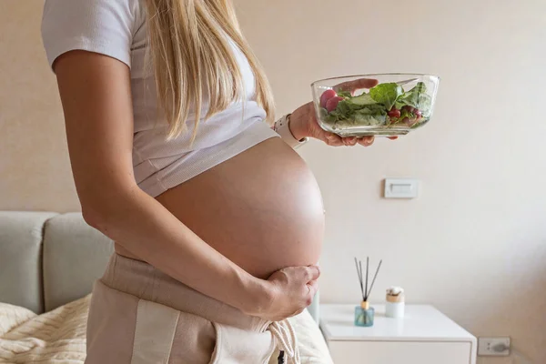 Feliz joven embarazada comiendo ensalada de verduras en casa. Concepto de nutrición saludable y embarazo. Esperando madre sosteniendo tazón con pepinos frescos y tomates —  Fotos de Stock