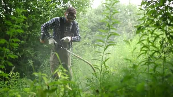 Человек в очках стрижет высокую пышную траву с триммером в саду — стоковое видео
