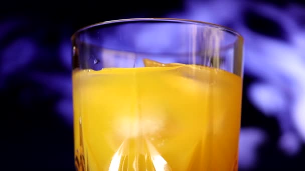 黄色とオレンジの甘いマルチフルーツジュースとアイスキューブでゆっくりとガラスを回転させます 色付きの煙を背景にジュースを塗ったグラス — ストック動画