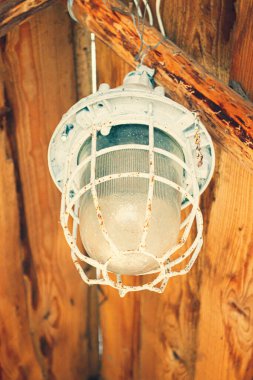 Çardağın ahşap tavanının altında eski bir fener. Gri-mavi boyayla boyanmış paslı metal fener. Cam platolu fener..