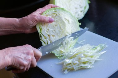 Beyaz bir kesim tahtasında, siyah bir masada taze lahana kesen yaşlı bir kadının elleri.