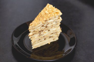 Kremalı bir parça puf böreği siyah bir seramik tabakta, pastanın tepesine kırıntılar serpiştirilmiş, katmanlar arasında kremayı görebilirsiniz. 