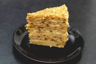 Kremalı bir parça puf böreği siyah bir seramik tabakta, pastanın tepesine kırıntılar serpiştirilmiş, katmanlar arasında kremayı görebilirsiniz. 