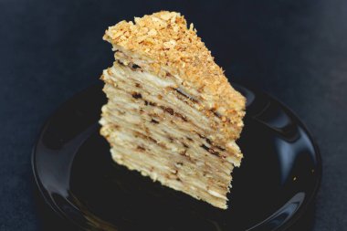 Kremalı bir parça puf böreği siyah bir seramik tabakta, pastanın tepesine kırıntılar serpiştirilmiş, katmanlar arasında kremayı görebilirsiniz. 