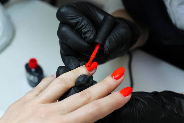 Manicure specialist in black gloves cares about hands nails. Manicurist paints nails with red nail polish