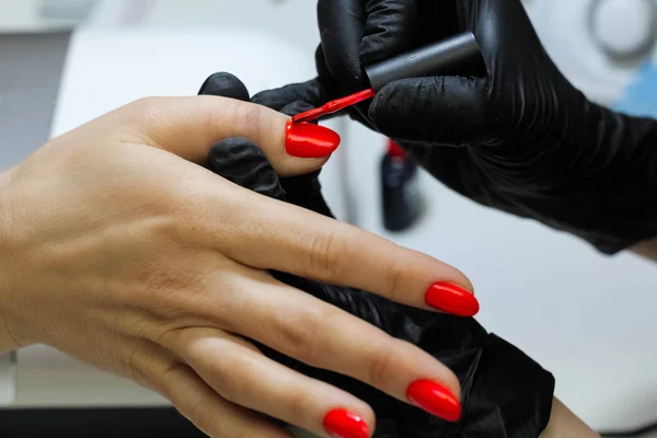Manicure specialist in black gloves cares about hands nails. Manicurist paints nails with red nail polish