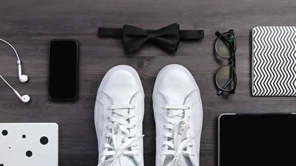 Modern men fashion accessories and electronic devices on dark background. White sneakers, tablet and phone flat lay