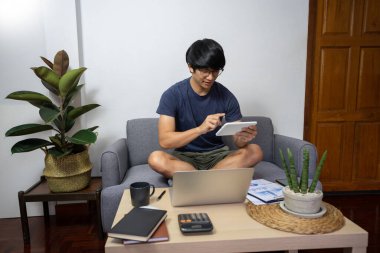 Evinde dizüstü bilgisayarıyla çalışan Asyalı bir girişimci. Evrak işleri ve belgeler oturma odasında, ev konseptinden çalışıyor.