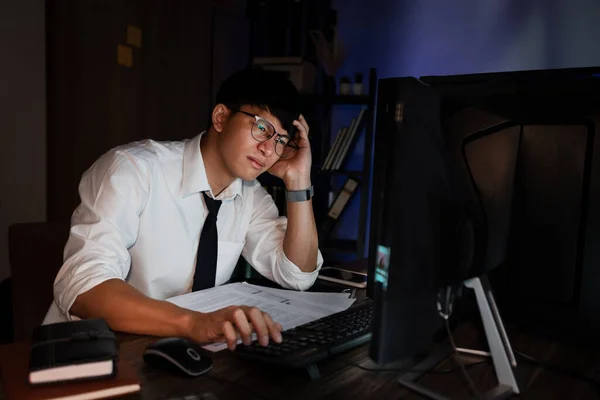 压力很大的年轻亚洲商人一个人在办公室工作到很晚 他的眼睛都要闭上了 看起来很头疼 加班超负荷工作的概念 — 图库照片