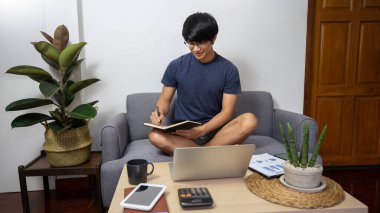 Evinden uzakta dizüstü bilgisayarıyla çalışan yakışıklı Asyalı serbest yazar, evdeki rahat bej koltukta oturuyor, günlük kıyafetlerini giyiyor.