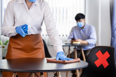 Garson, Coronavirus salgını için dezenfektan alkol şişesiyle (Covid-19) bir restoranda eldiven, yüz kalkanı ve koruyucu maske temizleme masası yüzeyi (prezervatif) takıyor..