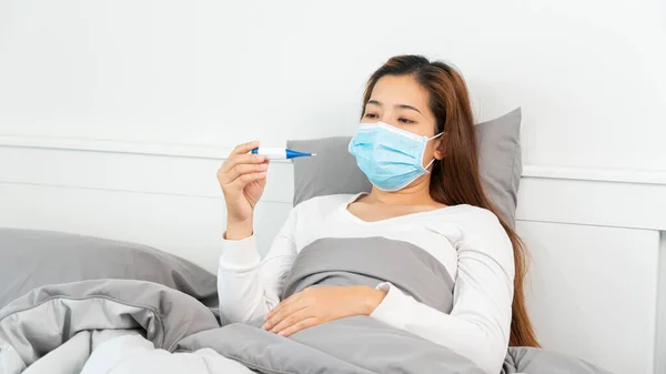 Sick Asian woman with protective mask lying in bed with high fever and measuring body temperature by thermometer and touching her forehead at her home