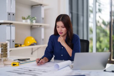 Odaklanmış Asyalı mimar kadın titizlikle bina planları projesini gözden geçiriyor modern ofis çalışma istasyonu tasarlıyor. Profesyonel kadın mühendis şehir gelişimini planlıyor.