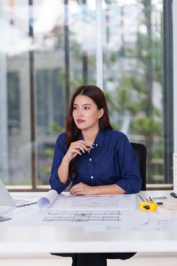 Profesyonel Asyalı kadın mimar parlak bir ofis masasında karmaşık planlar tasarlıyor. Yenilikçi vizyon ve stratejik planlama ile gelecekteki yapıları geliştiriyor..