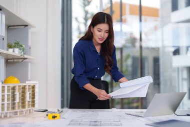 Odaklanmış Asyalı kadın mimar inşaat planları ve detaylı yapı modelleri üzerinde çalışıyor. Modern çalışma alanı yenilikçi bir tasarım yaratıyor.