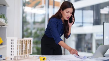 Asyalı yetişkin profesyonel mimar tasarım konseptlerini telefonla iletir. Bir dizüstü bilgisayarın ve bina modelinin proje planlarını inceliyor..