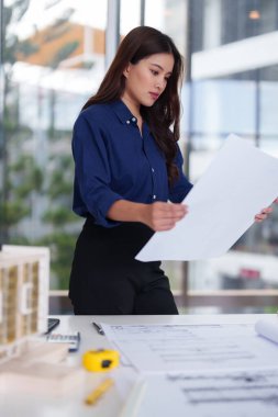 Odaklanmış Asyalı kadın mimar, mimari çizimleri özenle inceliyor. İnşaat projeleri için kentsel tasarım kavramları üzerinde çalışıyor..