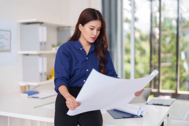 Çalışkan Asyalı kadın mimar, parlak modern ofis tasarım stüdyosunun ayrıntılı yapı planlarını analiz ediyor.