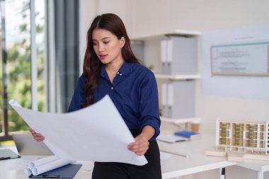 Profesyonel Asyalı kadın mimar, yapı ve proje tasarımına adanmış modern bir ofis stüdyosunun planlarını analiz ediyor.