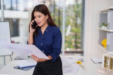 Yetişkin Asyalı kadın mimar müşteri projesinin detaylarını aktarıyor. Tasarım planlarını parlak profesyonel ofiste tutuyor.