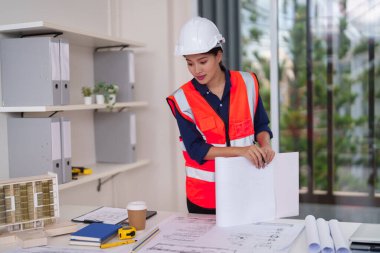 Profesyonel Asyalı mimar kadın mühendis, çağdaş bir tasarım stüdyosundaki karmaşık yapı planlarını ve mimari modelleri dikkatle inceliyor..