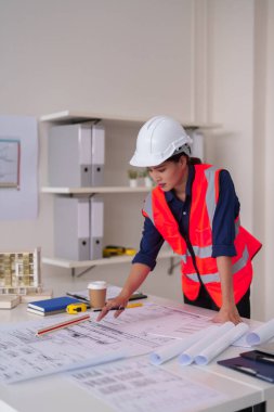 Çalışkan Asyalı kadın mimar güvenlik teçhizatı ile titizlikle modern bir ofis masasında yeni bir inşaat projesinin planlarını çalışıyor.
