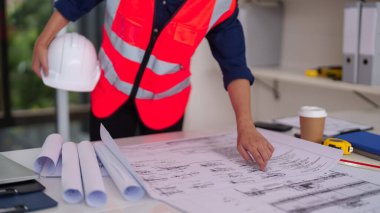 Asyalı mimar profesyonel incelemeler ofis masasındaki tasarım planları mühendislik inşaatı geliştirme kentsel planlaması için titiz proje analizleri yapıyor.