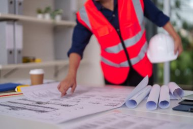Profesyonel mimar veya mühendis, modern ofis işyerindeki güvenlik ekipmanlarıyla birlikte masadaki ayrıntılı bina planlarını dikkatle inceler.