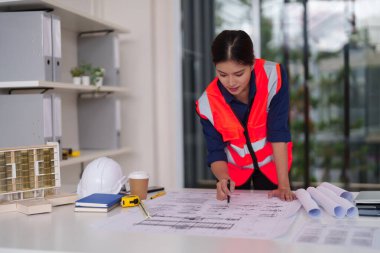 Modern bir ofisin kompleks bina planlarını inceleyen odaklanmış Asyalı kadın mimar. Onun profesyonel yaklaşımı projenin doğruluğunu ve yenilikçi tasarım çözümlerini güvence altına alıyor.