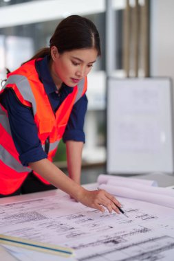 Modern bir ofisin mimari planlarını inceleyen Asyalı kadın mühendis. Uzmanlık alanı proje planlama üzerine..
