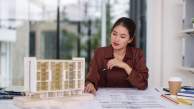 Asyalı kadın mimar yoğun bir şekilde bina prototipi ve detaylı inşaat planları üzerinde çalışıyor. Onun profesyonel adanmışlığı yenilikçi tasarımı ve gelecekteki şehir gelişimini yönlendiriyor..