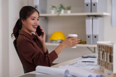 Asyalı kadın mimar gülümsüyor akıllı telefonla konuşuyor canlı tasarım stüdyosunda elinde kahve fincanı, planlar ve bina modeli ile.