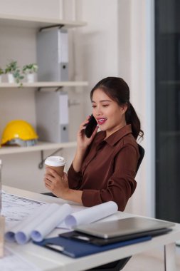 Yetişkin Asyalı mimar proje detaylarını verimli bir ofis ortamında mobil iletişim yoluyla tartışır. Elinde sıcak bir içecek var. Profesyonel çalışma alanı, kariyer odaklanma, yenilik.